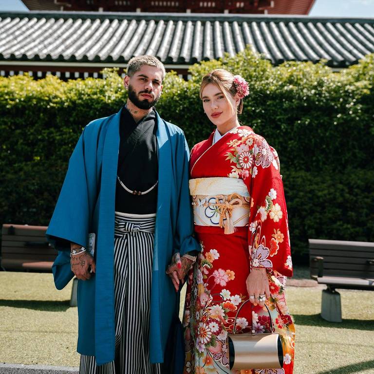 Um casal posando em frente a um templo japonês. O homem está vestindo um kimono azul com calças listradas, enquanto a mulher usa um quimono vermelho com estampas florais. Ambos estão segurando as mãos e sorrindo. Ao fundo, pode-se ver a estrutura do templo com telhados vermelhos e detalhes arquitetônicos tradicionais.