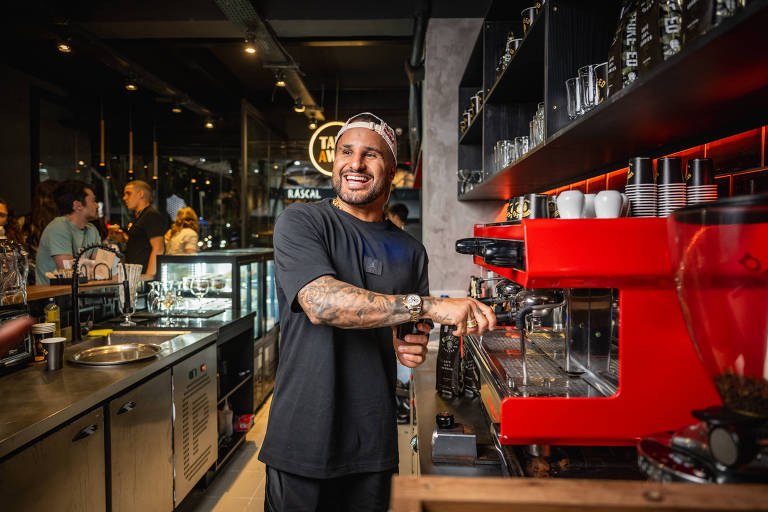 Um barista sorridente está posicionado em frente a uma máquina de café vermelha em um café moderno. Ele usa uma camiseta preta e um boné. O ambiente é iluminado e há várias pessoas ao fundo, além de utensílios de café e uma vitrine com bebidas. O barista parece estar preparando uma bebida, com um sorriso no rosto.