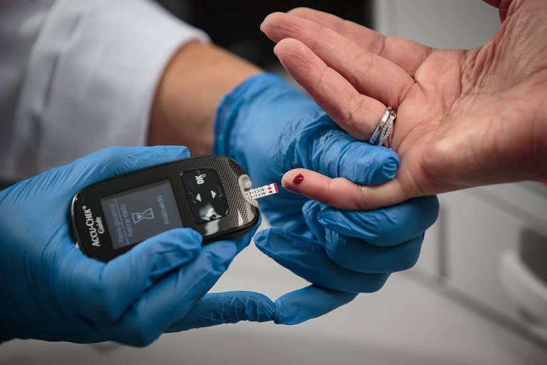 Uma pessoa está realizando um teste de glicemia. A mão da pessoa que está sendo testada é segurada por outra mão que usa luvas azuis. Um dispositivo de medição de glicemia está sendo colocado na ponta do dedo, onde há uma gota de sangue. O fundo é desfocado, mas parece ser um ambiente clínico.