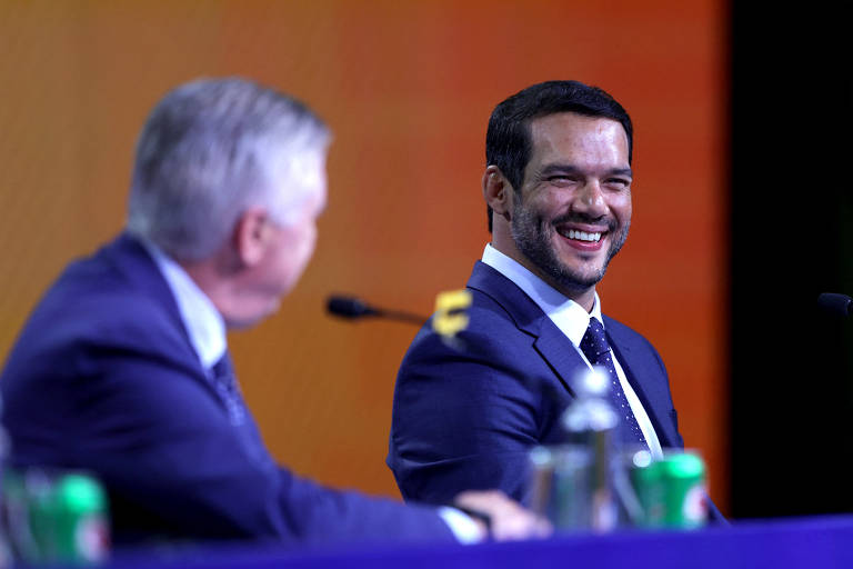 A imagem mostra dois homens em um evento de conferência. O homem à esquerda, com cabelo grisalho e terno escuro, está olhando para o homem à direita, que está sorrindo e usando um terno azul. O fundo é colorido, com tons de laranja e verde, e há garrafas de refrigerante em uma mesa à frente.