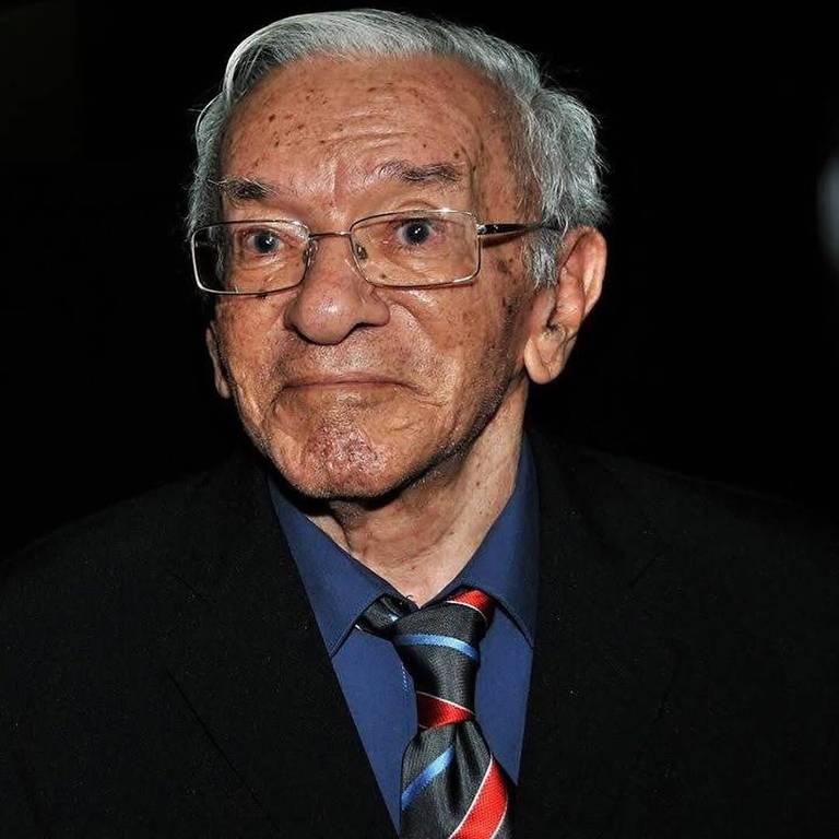 A imagem mostra um homem idoso com cabelo grisalho e pele clara, usando óculos. Ele está vestido com uma camisa azul e um paletó escuro, além de uma gravata com listras vermelhas e azuis. O fundo da imagem é escuro, destacando o rosto do homem, que tem uma expressão séria.