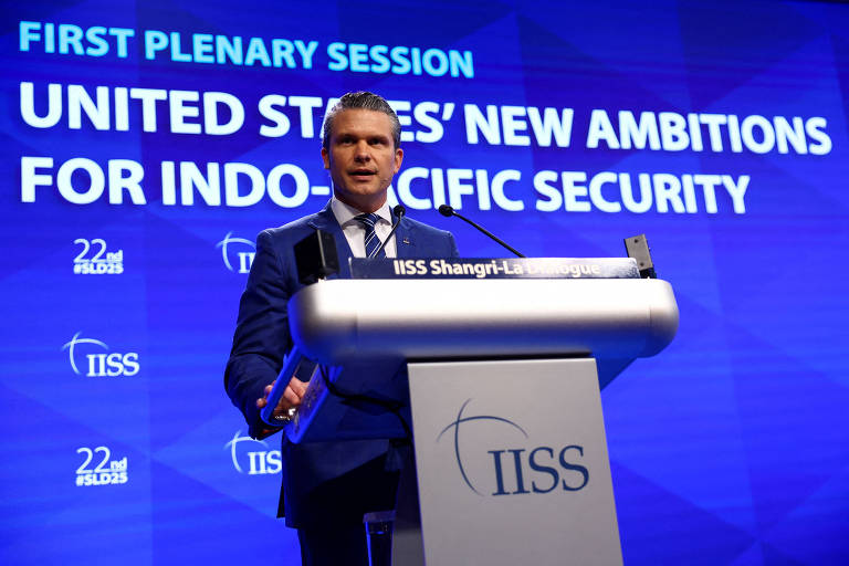 Pete Hegseth, um homem em um terno azul está falando em um púlpito durante uma sessão plenária. Ao fundo, há um grande painel com o texto "FIRST PLENARY SESSION UNITED STATES' NEW AMBITIONS FOR INDO-PACIFIC SECURITY". O evento parece ser uma conferência, com o logotipo do IISS visível no púlpito e no fundo. A atmosfera é formal e profissional.