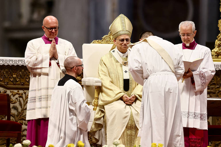 A imagem mostra uma cerimônia religiosa em um ambiente grandioso. No centro, um homem vestido com vestes litúrgicas, incluindo um manto dourado e um chapéu de bispo, está sentado em uma cadeira. À sua frente, um outro homem, também vestido de branco, está se ajoelhando e fazendo uma reverência. Ao fundo, há outros clérigos, um deles de óculos e com cabelo raspado, e outro com vestes roxas, que observam a cena. O ambiente é decorado com flores amarelas na frente.