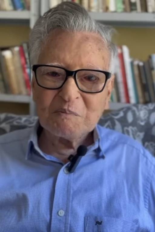 Um homem idoso está sentado em um sofá, usando uma camisa azul e óculos. Ele tem cabelo grisalho e uma expressão serena. Ao fundo, há uma estante com livros organizados. A iluminação é suave, criando um ambiente acolhedor.