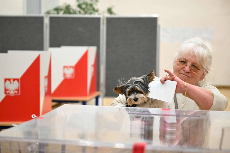 Uma mulher idosa está votando em uma cabine eleitoral, segurando um papel de voto. Ao seu lado, um pequeno cachorro está olhando para a urna. Ao fundo, há várias cabines de votação com a bandeira da Polônia visível.