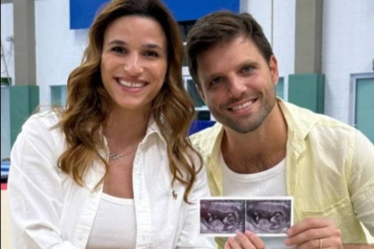 Um casal está sentado no chão, sorrindo e segurando cartões. A mulher tem cabelo longo e ondulado, vestindo uma blusa branca e uma saia clara. O homem tem cabelo curto e usa uma camiseta clara e calças jeans. Ao fundo, há um ambiente que parece ser uma instalação esportiva, com uma bandeira do Brasil visível na parede.