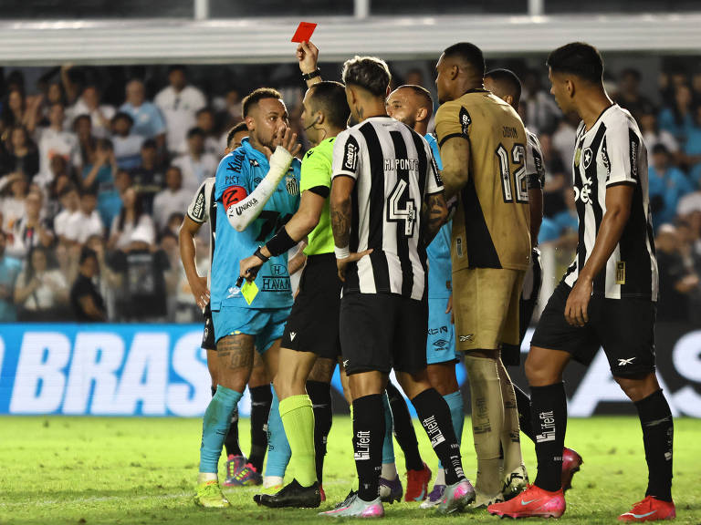 A imagem mostra um momento de tensão em um jogo de futebol, onde um árbitro está mostrando um cartão vermelho para um jogador. O jogador, que está usando um uniforme azul, parece estar discutindo com o árbitro, que está vestido de amarelo. Outros jogadores, de diferentes uniformes, estão ao redor, observando a situação. O fundo da imagem mostra uma multidão de torcedores.