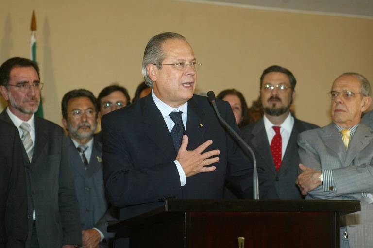 Um homem em um terno escuro está falando em um microfone, com a mão sobre o peito, em um evento oficial. Atrás dele, há várias pessoas, algumas com ternos e gravatas, observando atentamente. Ao fundo, uma bandeira do Brasil é visível. O ambiente parece ser uma sala de conferências ou um auditório.