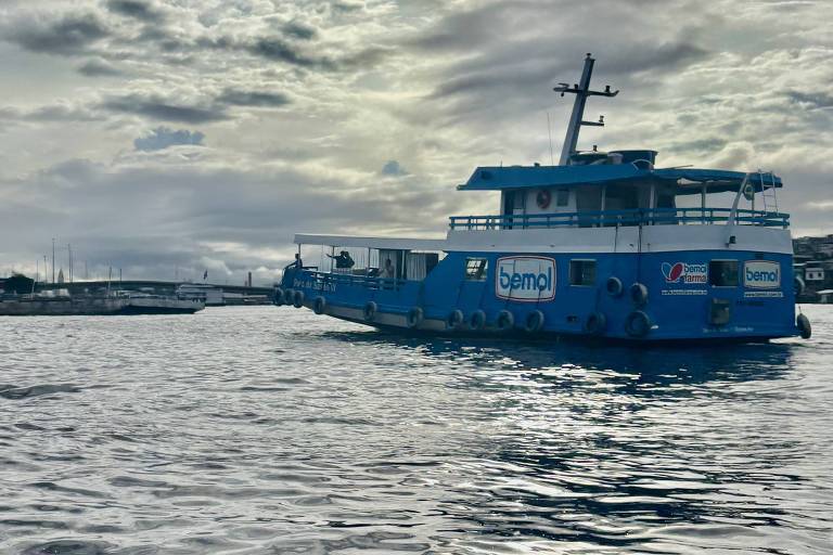 A imagem mostra um barco azul navegando em águas calmas. O céu está nublado, com nuvens escuras e claras. O barco possui o nome 'Semo' visível em sua lateral e parece estar se aproximando de um cais ao fundo. A superfície da água reflete a luz do céu, criando um efeito brilhante.