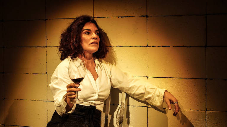 A imagem mostra uma mulher sentada em uma cadeira, segurando uma taça de vinho tinto. Ela tem cabelos cacheados e escuros, e usa uma camisa branca. O fundo é uma parede de blocos de concreto, iluminada de forma suave, criando um contraste com a figura da mulher. A expressão dela parece contemplativa ou pensativa.