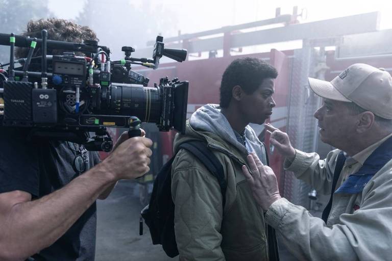 A imagem mostra uma cena de filmagem com um cinegrafista segurando uma câmera profissional voltada para um ator. O ator, que está usando um casaco, parece estar em uma conversa com um homem mais velho, que está ajustando algo em seu rosto. O ambiente é nebuloso, com estruturas metálicas ao fundo, sugerindo um set de filmagem. Há também um logotipo da Paramount+ no canto inferior direito.