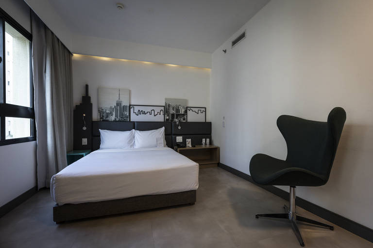 Um quarto de hotel moderno com uma cama de casal, lençóis brancos e almofadas. Ao fundo, há uma parede com uma decoração que inclui uma imagem de uma cidade. O quarto possui uma janela grande com cortinas cinzas e um piso de cerâmica. No canto, há uma cadeira estilizada em formato de concha, de cor verde escura, e uma mesa de cabeceira ao lado da cama