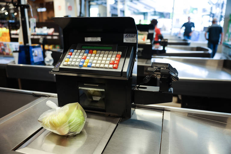 A imagem mostra uma caixa registradora em um supermercado. A caixa é preta e possui um teclado colorido com botões de diferentes cores. Na frente da caixa, há uma sacola plástica com maçãs verdes. Ao fundo, é possível ver outras pessoas e prateleiras de produtos, indicando que o ambiente é um supermercado.
