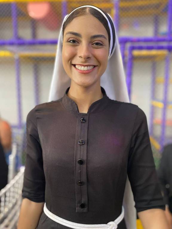 Uma mulher sorridente está vestida com um traje tradicional escuro, que possui botões na parte frontal e uma faixa branca na cintura. Ela usa um lenço branco na cabeça e está em um ambiente que parece ser uma área de recreação, com estruturas coloridas ao fundo. A mulher está em pé, com as mãos na cintura, e parece estar em um momento de descontração.