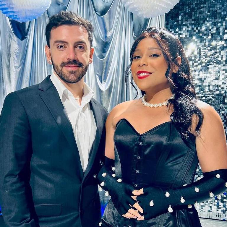 Um homem e uma mulher posam juntos em um cenário de evento formal. O homem está vestido com um terno preto e uma camisa branca, enquanto a mulher usa um vestido preto elegante com detalhes brilhantes e luvas longas. O fundo é decorado com cortinas prateadas e esferas brancas, criando uma atmosfera festiva.