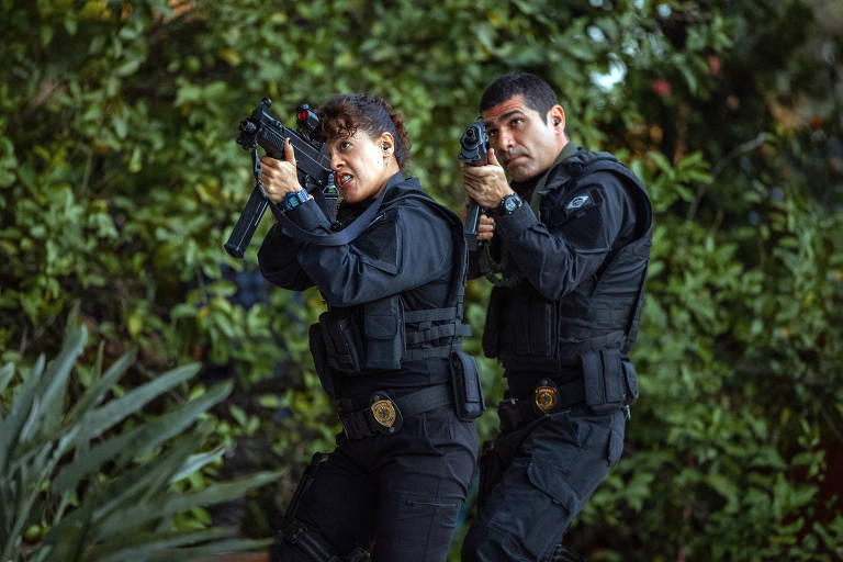 A imagem mostra dois policiais em uma posição de ataque, ambos armados. A policial à esquerda segura uma submetralhadora e tem uma expressão intensa, enquanto o policial à direita está com uma pistola. Ambos estão vestidos com uniformes pretos e coletes táticos, e há vegetação ao fundo, sugerindo um ambiente externo.