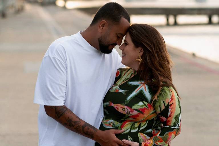 Um casal está se abraçando em um ambiente ao ar livre, com um porto ao fundo. O homem usa uma camiseta branca e a mulher está vestida com um vestido colorido com estampas de folhas. Eles estão sorrindo um para o outro, com a mulher segurando a barriga, indicando uma gravidez. A luz do sol está suave, criando uma atmosfera romântica.