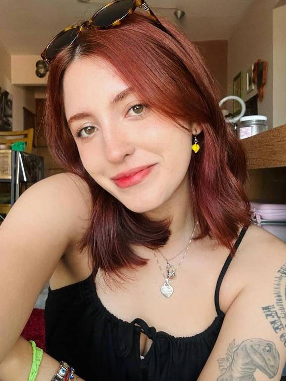 A imagem mostra uma jovem mulher com cabelo vermelho e liso, usando uma blusa preta. Ela está sorrindo e posando para a câmera, com um braço estendido para frente. A mulher tem vários tatuagens visíveis nos braços e usa acessórios coloridos, como brincos e pulseiras. Ao fundo, é possível ver uma sala de estar com móveis e decoração variados, incluindo um sofá e prateleiras.