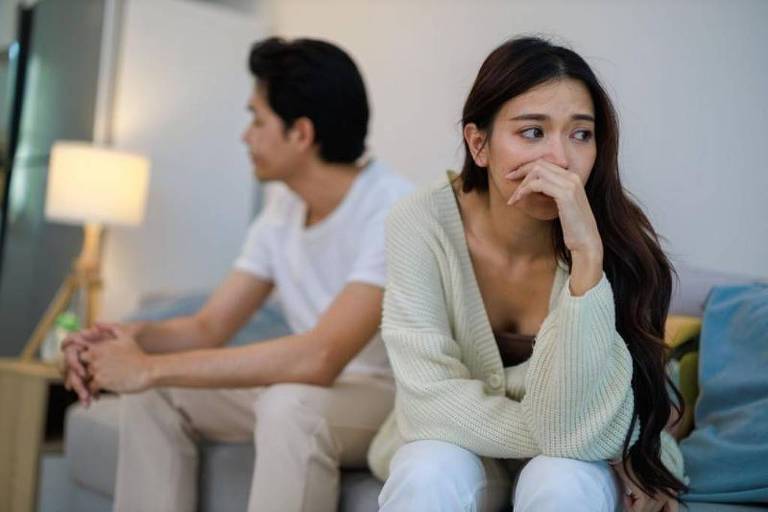 A imagem mostra um casal sentado em um sofá, ambos aparentando estar em desacordo. A mulher, com cabelo longo e solto, está olhando para baixo com uma expressão preocupada, enquanto o homem, de costas, parece distante e pensativo. O ambiente é moderno, com uma luz suave ao fundo e almofadas coloridas no sofá.