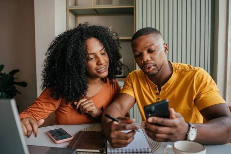 A imagem mostra um casal sentado em uma mesa. A mulher, com cabelo cacheado e vestido laranja, está olhando para o homem ao seu lado, que está usando uma camiseta amarela e segurando um smartphone. Ele parece estar explicando algo enquanto escreve em um caderno. Na mesa, há um laptop, um bloco de notas e uma caneca. O ambiente é bem iluminado e moderno, com prateleiras ao fundo.