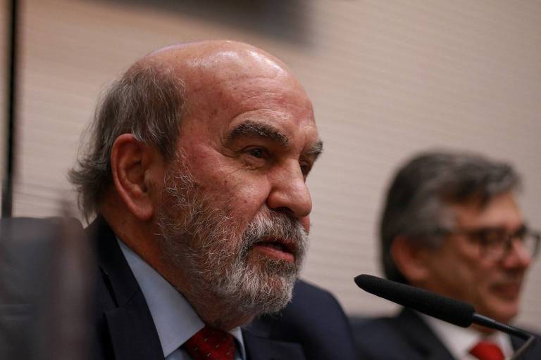 A imagem mostra um homem de barba e cabelo grisalho, usando um terno escuro e uma gravata vermelha, falando em um microfone. Ao fundo, há outro homem com óculos e cabelo grisalho, que parece estar ouvindo atentamente. O ambiente é formal, com uma parede clara ao fundo.