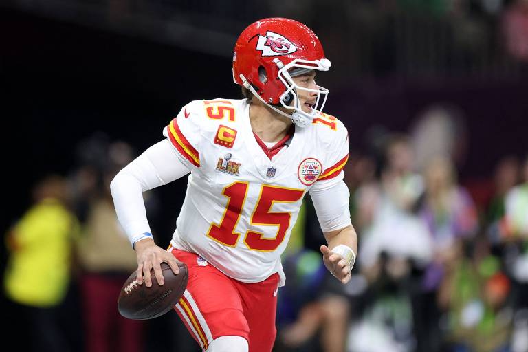Um jogador de futebol americano está em movimento, lançando uma bola. Ele veste um uniforme vermelho e branco, com o número 15 nas costas. O jogador tem um capacete vermelho e está em um campo de jogo, com torcedores ao fundo. A imagem captura um momento dinâmico do jogo.
