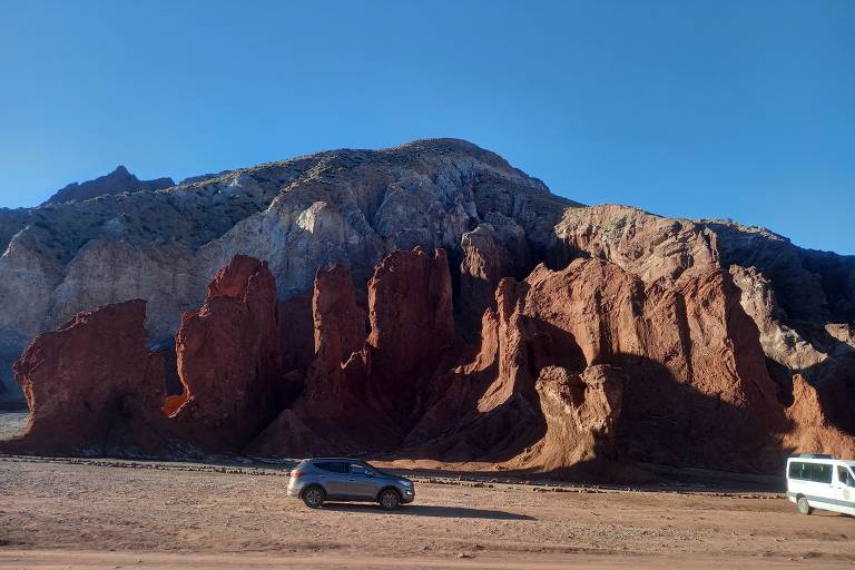 A imagem mostra uma paisagem desértica com formações rochosas de cores avermelhadas e marrons. No primeiro plano, há um carro cinza passando ao lado das rochas, enquanto ao fundo se vê uma montanha com uma coloração mais clara. O céu está limpo e azul, indicando um dia ensolarado.