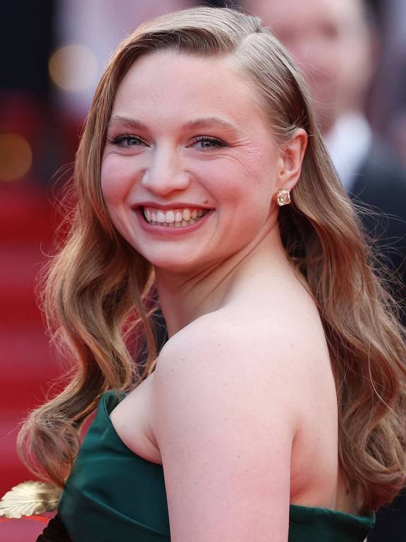 Uma mulher sorridente com cabelo longo e ondulado, vestindo um vestido verde. Ela está em um evento de tapete vermelho, com um fundo desfocado de pessoas e luzes. A mulher parece estar posando para a câmera, com uma expressão alegre e confiante.