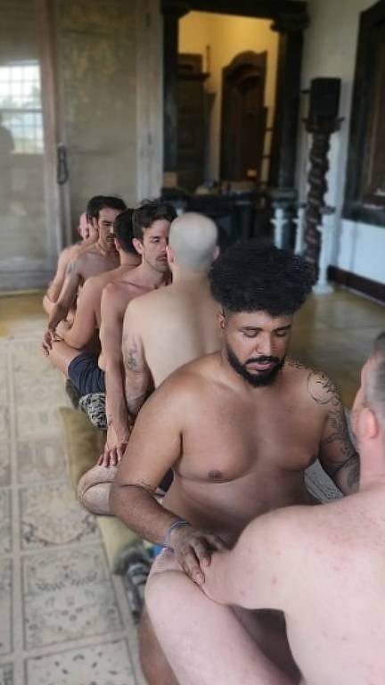 A imagem mostra um grupo de seis homens sentados em uma fila, todos sem roupas, em um ambiente interno. Eles estão posicionados de costas uns para os outros, com um homem na frente realizando um gesto de massagem nas costas do homem à sua frente. O ambiente parece ser uma sala com paredes claras e móveis de madeira ao fundo.