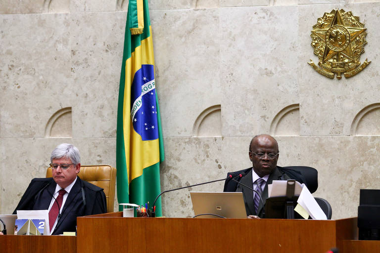 A imagem mostra uma sessão do Supremo Tribunal Federal do Brasil. À esquerda, um homem de terno e gravata está sentado em uma cadeira, enquanto à direita, outro homem, também de terno, está falando ou apresentando algo. Ao fundo, há uma bandeira do Brasil e o brasão da República. O ambiente é formal e austero, típico de uma corte judicial.