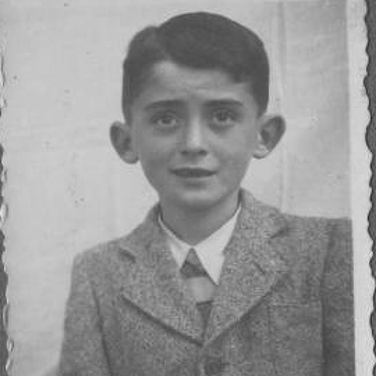 A imagem é uma fotografia em preto e branco de um menino. Ele está usando um terno cinza claro com um colete e uma gravata. O menino tem cabelo escuro e liso, e está olhando diretamente para a câmera com uma expressão neutra. O fundo da imagem é claro e a foto tem bordas recortadas.