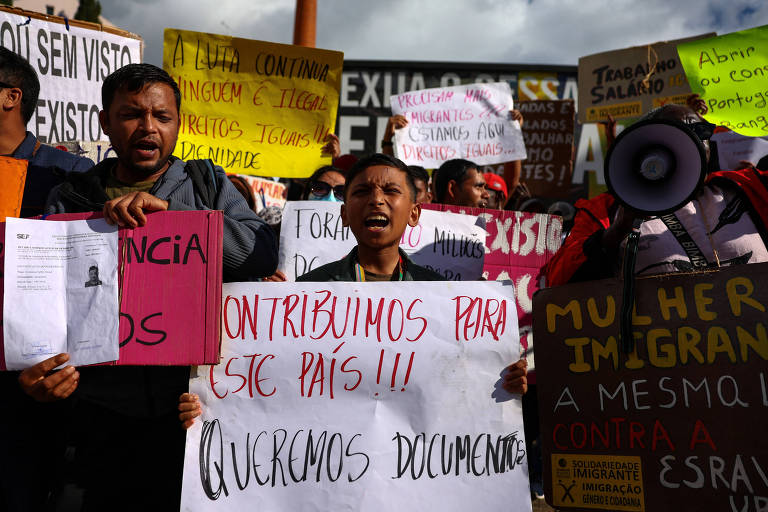 A imagem mostra um grupo de pessoas participando de um protesto. Algumas pessoas estão segurando cartazes e faixas com mensagens sobre direitos e imigração. Um dos cartazes diz 'CONTRIBUÍMOS PARA ESTE PAÍS!!!' e outro menciona 'QUEREMOS DOCUMENTOS'. O ambiente parece ser ao ar livre, com um céu parcialmente nublado ao fundo.