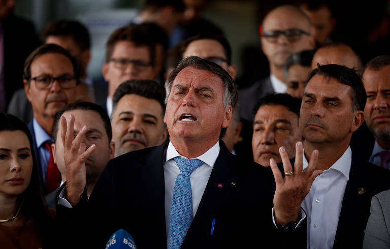 A imagem mostra um grupo de homens e mulheres em uma coletiva de imprensa. Um homem em destaque, com cabelo grisalho e terno escuro, está gesticulando com as mãos enquanto fala. Ele usa uma gravata azul e parece estar expressando uma opinião forte. Ao seu redor, há várias pessoas, algumas com expressões sérias, que o escutam atentamente.