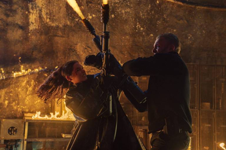 A imagem mostra uma cena de ação em um ambiente escuro e flamejante. Uma mulher com cabelo longo e solto está lutando contra um homem, ambos segurando uma arma. O homem parece estar tentando desarmar a mulher, enquanto ela se esforça para manter o controle da arma. Ao fundo, há chamas e uma estrutura metálica, sugerindo um cenário de combate intenso.