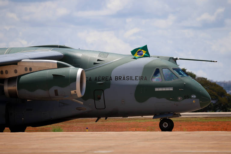 A imagem mostra a parte frontal de um avião da Força Aérea Brasileira, com uma pintura camuflada em tons de verde. Na parte superior do avião, há uma bandeira do Brasil. O fundo é um céu parcialmente nublado e uma pista de pouso.