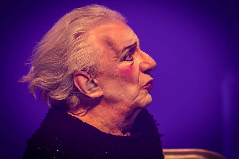 A imagem mostra um homem idoso com cabelo grisalho e liso, usando maquiagem de palhaço, incluindo blush e batom. Ele está vestido com um cachecol preto e tem uma expressão pensativa. O fundo é de cor roxa, criando um contraste com o rosto iluminado do homem.