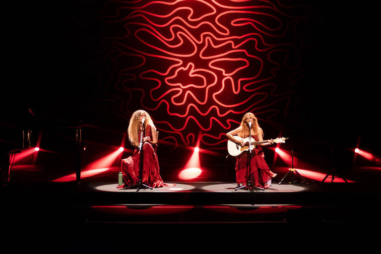 Duas mulheres estão se apresentando em um palco. Uma delas está tocando violão, enquanto a outra canta. Ambas estão vestidas com longos vestidos vermelhos. O fundo do palco possui uma iluminação dramática com um padrão ondulado em vermelho e laranja, criando um ambiente vibrante. Há também algumas luzes direcionais que iluminam as artistas.