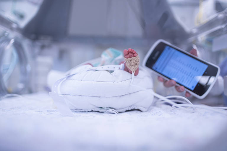 A imagem mostra um bebê em uma incubadora, com um pé visível e um dispositivo de monitoramento conectado. Ao lado do bebê, há um smartphone que exibe uma tela com informações, possivelmente relacionadas à saúde do bebê. O ambiente parece ser uma unidade de terapia intensiva neonatal, com equipamentos médicos ao fundo.