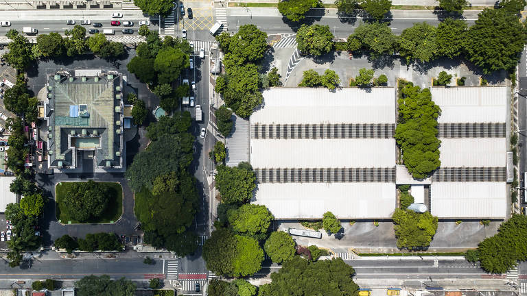 A imagem mostra uma vista aérea de uma área urbana. À esquerda, há um edifício com um telhado verde e cercado por árvores. À direita, há um grande edifício com telhado claro, também cercado por vegetação. A área ao redor é predominantemente coberta por árvores, e há ruas visíveis na parte inferior da imagem.
