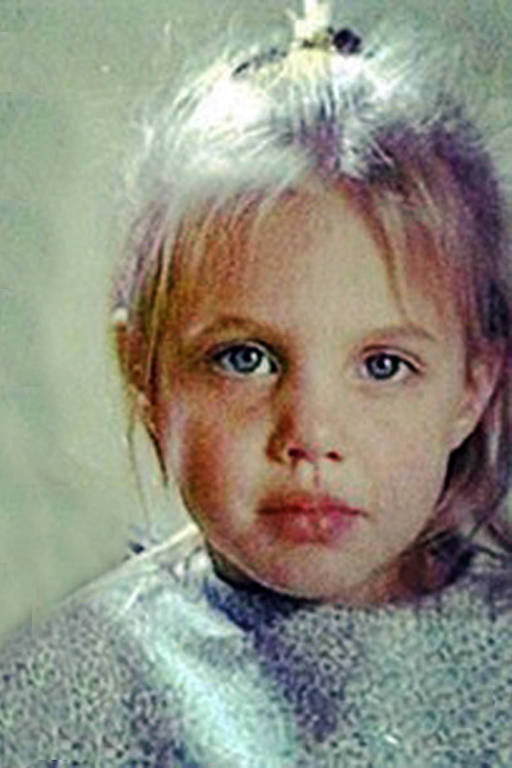 A imagem mostra o rosto de uma criança com cabelo loiro e liso, preso em um coque. Ela tem olhos azuis e uma expressão séria. A criança está vestindo uma blusa de cor clara com um padrão sutil. O fundo da imagem é desfocado, dando destaque ao rosto da criança.