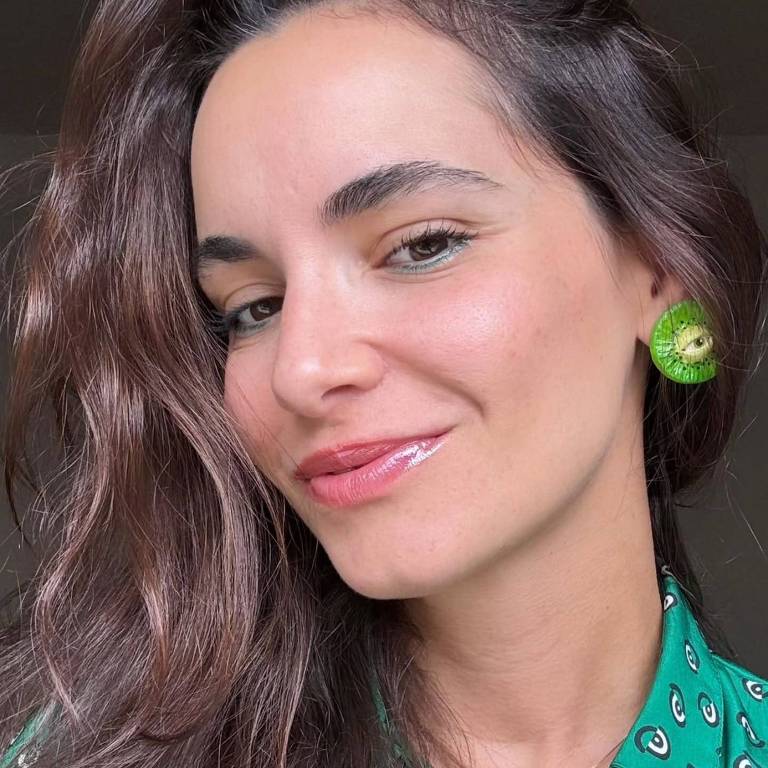 A imagem mostra uma mulher sorrindo, com cabelo longo e ondulado. Ela está usando uma blusa verde com padrões e brincos grandes e verdes. O fundo é neutro, destacando seu rosto e a blusa.