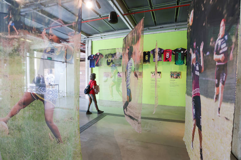 A imagem mostra uma exposição interna com painéis de fotos transparentes pendurados, retratando atletas em ação. Ao fundo, há uma parede verde com camisas de esporte expostas. Uma pessoa está caminhando pelo espaço, passando entre os painéis de fotos.