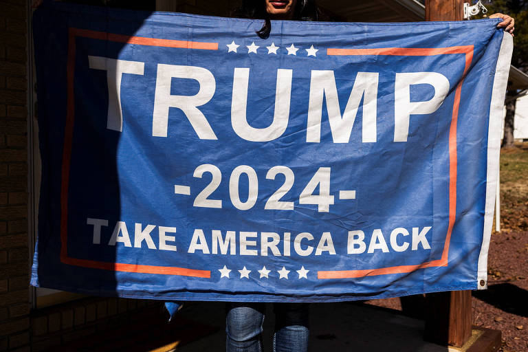 Uma pessoa está segurando uma bandeira azul com o texto em letras grandes: 'TRUMP - 2024' e abaixo, 'TAKE AMERICA BACK'. A bandeira tem um design com estrelas brancas e listras ao redor do texto.