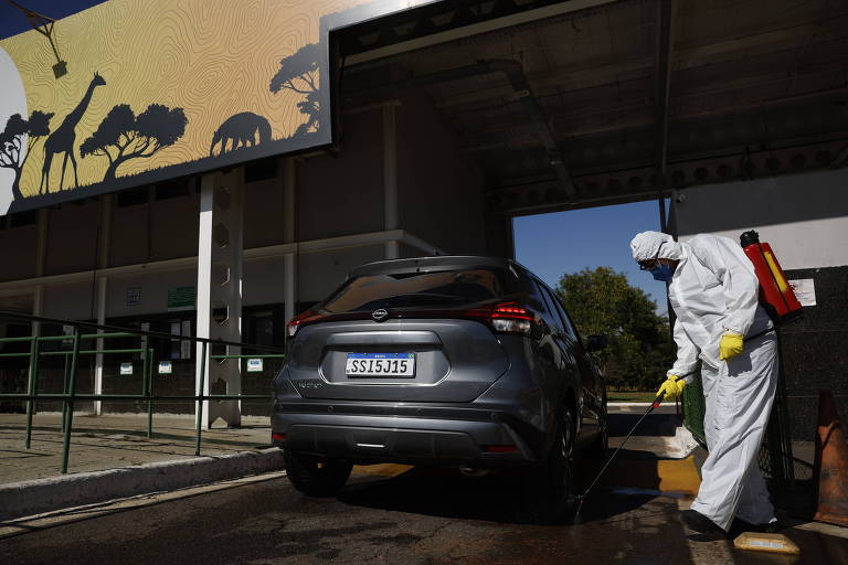 Een arbeider in een beschermend pak en handschoenen desinfecteert een grijze auto in een wasstraat. De auto staat geparkeerd in een rijstrook en de arbeider gebruikt een spuitpistool. Op de achtergrond is een paneel met silhouetten van dieren en bomen te zien, wat een natuurlijke omgeving suggereert.