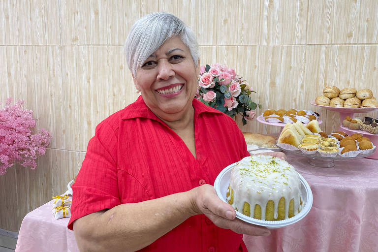 Uma mulher sorridente com cabelo curto e grisalho está segurando um bolo decorado com cobertura branca e raspas de limão. Ela usa uma camisa vermelha e está em um ambiente decorado com flores e uma mesa cheia de doces e salgados ao fundo.