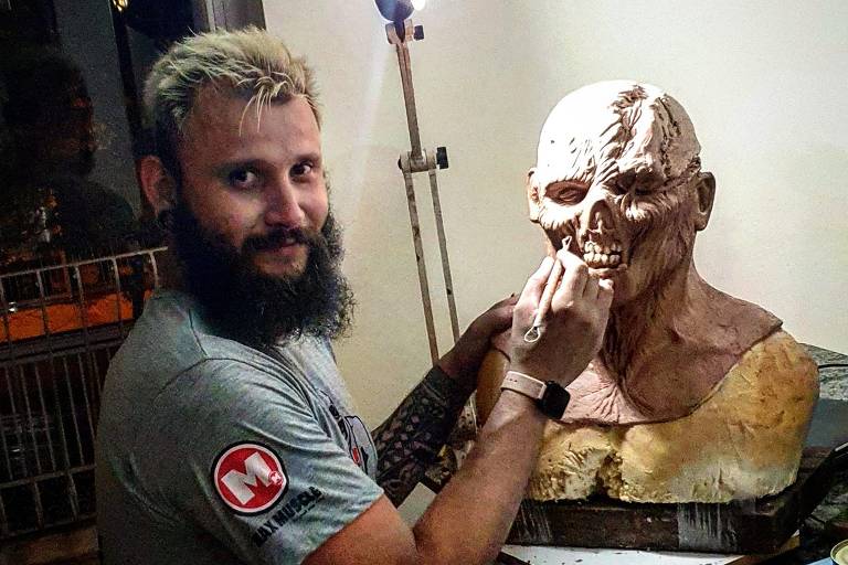 Um artista com barba está trabalhando em uma escultura de um rosto humano. Ele está usando ferramentas para detalhar a escultura, que parece ser feita de um material semelhante a argila ou cera. O ambiente é iluminado por uma luz artificial, e ao fundo, há uma estrutura que parece ser uma grade ou um suporte. O artista veste uma camiseta cinza e tem um adesivo ou emblema na manga.