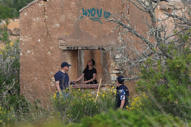 A imagem mostra três pessoas em uma área externa, próximas a uma construção em ruínas. A estrutura é de cor marrom e apresenta paredes desgastadas, com grafite visível na parte superior. Duas pessoas estão em pé, uma delas é um homem usando um boné e a outra é uma mulher que parece estar interagindo com a estrutura. A terceira pessoa, um homem com uniforme, está mais distante, observando. O ambiente ao redor é verde, com vegetação e flores.