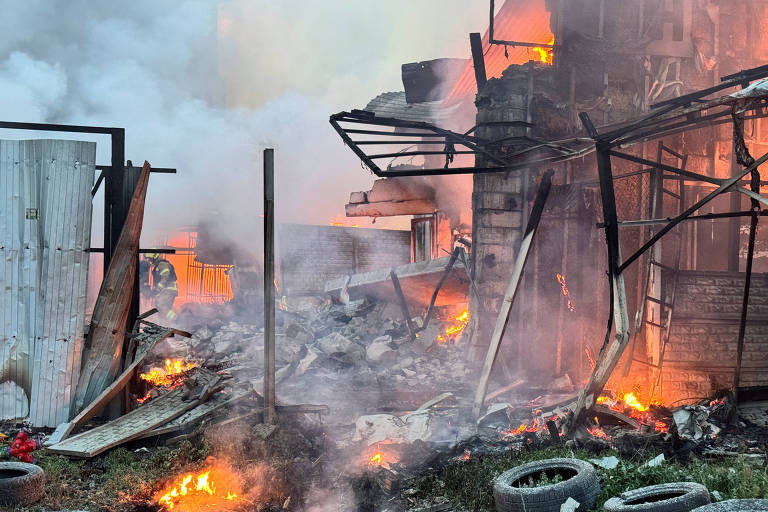 A imagem mostra uma cena de incêndio em uma estrutura, com chamas visíveis e fumaça densa. O local parece estar em ruínas, com destroços espalhados pelo chão e pneus queimando ao redor. A fumaça se eleva em direção ao céu, criando um ambiente caótico e perigoso.