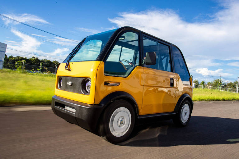 Um carro elétrico compacto e moderno, pintado de amarelo, está em movimento em uma estrada. O veículo possui um design distinto com janelas grandes e uma forma arredondada. O fundo apresenta um campo verde e um céu azul com algumas nuvens.