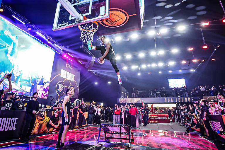 A imagem mostra um jogador de basquete realizando uma enterrada em uma quadra iluminada, com uma grande audiência ao fundo. O jogador está no ar, segurando a bola com uma mão, enquanto a outra mão está na borda da cesta. Há várias telas e luzes ao redor, criando um ambiente vibrante e animado. Algumas pessoas na plateia estão assistindo e aplaudindo.